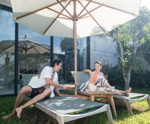 A man and woman relax on sun loungers under a large umbrella in a lush garden, sharing refreshments. The scene is calm and sunny, evoking a sense of leisure.