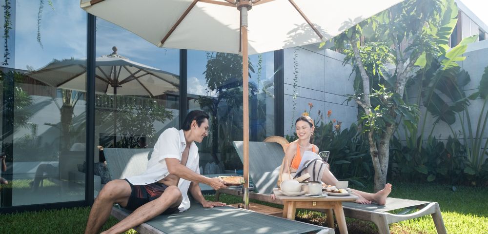 A man and woman relax on sun loungers under a large umbrella in a lush garden, sharing refreshments. The scene is calm and sunny, evoking a sense of leisure.