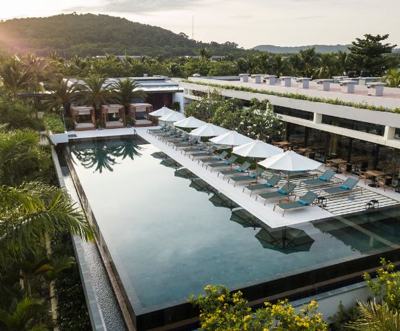 A tranquil hotel pool scene with rows of sunbeds and umbrellas beside a long, sleek pool. Surrounded by lush greenery and distant hills under a soft sky.