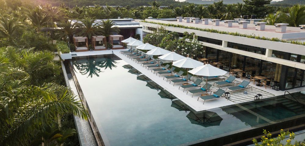 A tranquil hotel pool scene with rows of sunbeds and umbrellas beside a long, sleek pool. Surrounded by lush greenery and distant hills under a soft sky.