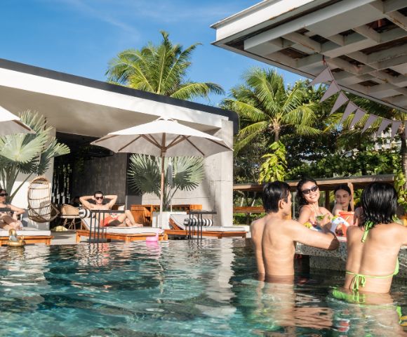 People enjoying a sunny day at a poolside bar, chatting and laughing. A couple relaxes on lounge chairs under umbrellas. Palm trees and blue sky in the background.