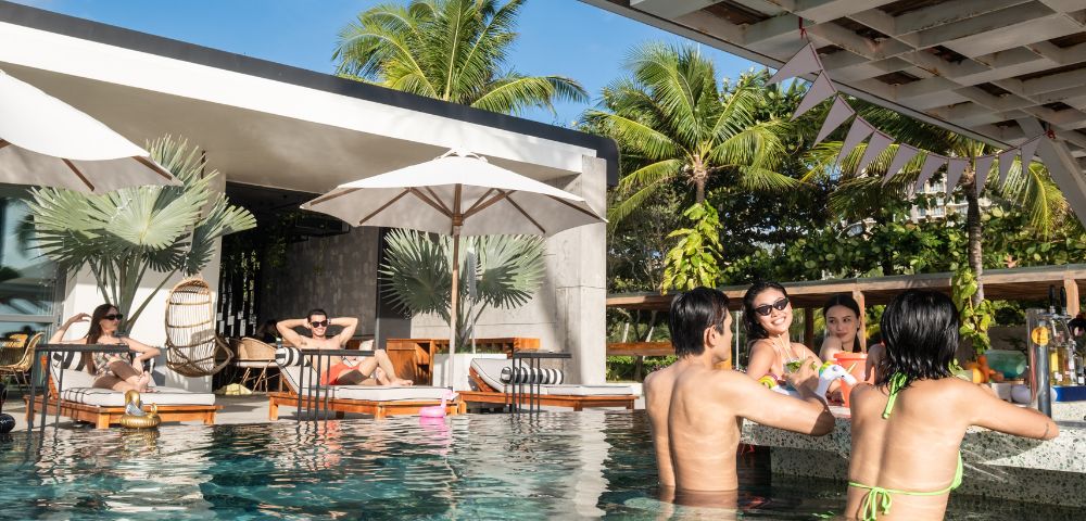 People enjoying a sunny day at a poolside bar, chatting and laughing. A couple relaxes on lounge chairs under umbrellas. Palm trees and blue sky in the background.