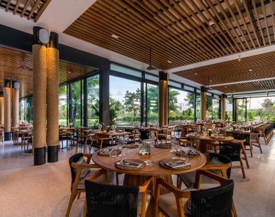 Modern restaurant interior with round wooden tables, neatly set with plates and glasses. Large windows offer views of lush greenery, creating a calm atmosphere.