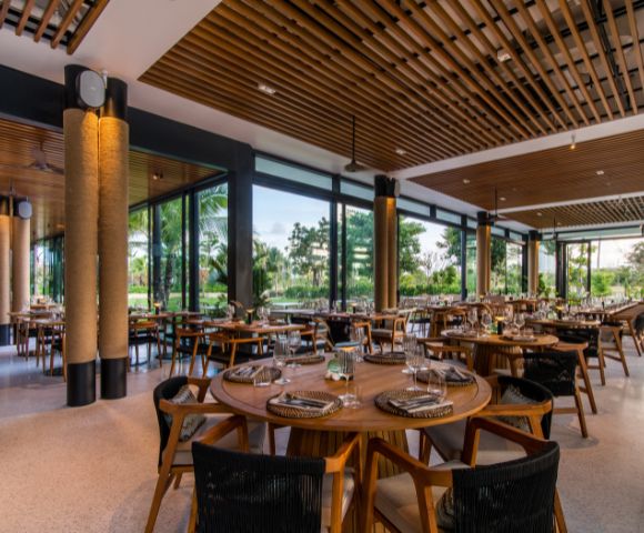 Modern restaurant interior with round wooden tables, neatly set with plates and glasses. Large windows offer views of lush greenery, creating a calm atmosphere.