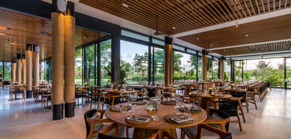Modern restaurant interior with round wooden tables, neatly set with plates and glasses. Large windows offer views of lush greenery, creating a calm atmosphere.