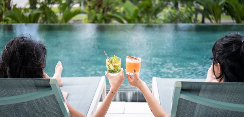 Two people relax on poolside loungers, holding colorful cocktails. The serene pool and lush greenery create a tranquil, tropical vibe.