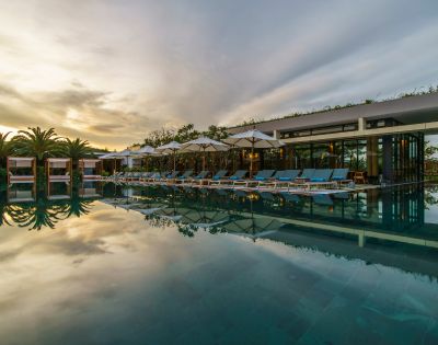 Luxurious poolside scene at sunset. Empty lounge chairs and umbrellas line the pool, reflecting on calm water. Palm trees and modern building in the background.