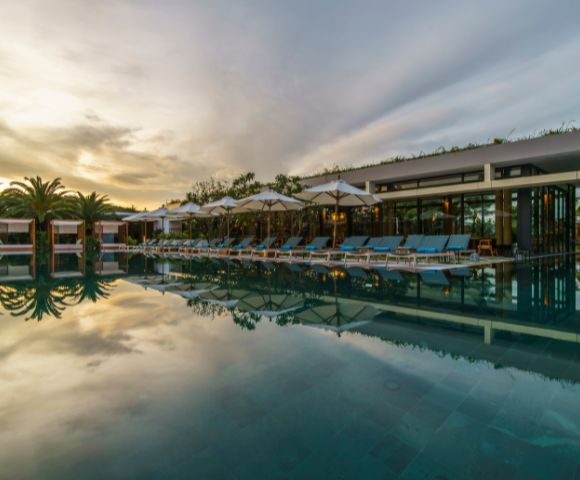 Luxurious poolside scene at sunset. Empty lounge chairs and umbrellas line the pool, reflecting on calm water. Palm trees and modern building in the background.