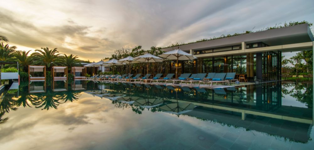 Luxurious poolside scene at sunset. Empty lounge chairs and umbrellas line the pool, reflecting on calm water. Palm trees and modern building in the background.