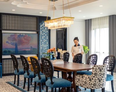 Elegant dining room with a long wooden table, blue-patterned chairs, chandelier, art on the wall, and a smiling woman setting the table, creating a warm, welcoming atmosphere.