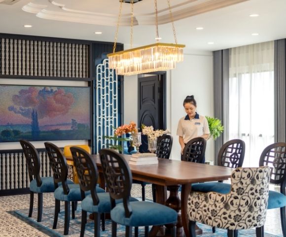 Elegant dining room with a long wooden table, blue-patterned chairs, chandelier, art on the wall, and a smiling woman setting the table, creating a warm, welcoming atmosphere.