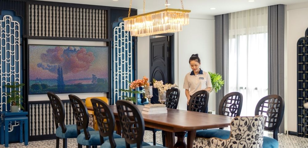 Elegant dining room with a long wooden table, blue-patterned chairs, chandelier, art on the wall, and a smiling woman setting the table, creating a warm, welcoming atmosphere.