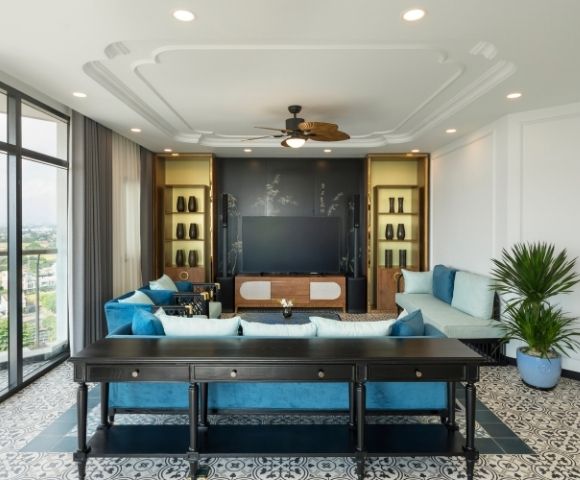A modern living room with blue and cream sofas, a dark wood console table, a large TV on a stand, patterned floor tiles, and a ceiling fan. A potted plant sits to the side. Natural light streams in from large windows on the left.