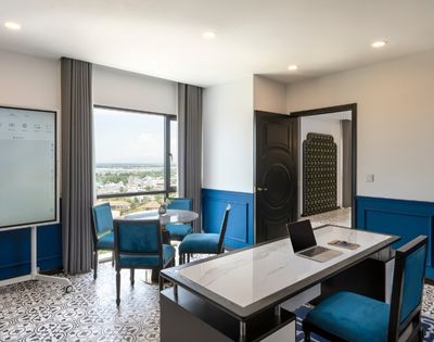 Modern office with a sleek desk, laptop, and four blue chairs around a round table. Large window offers scenic views. Blue accents create a calm atmosphere.