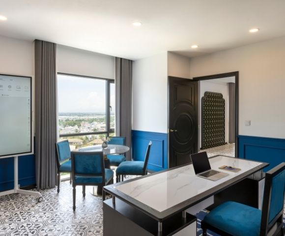 Modern office with a sleek desk, laptop, and four blue chairs around a round table. Large window offers scenic views. Blue accents create a calm atmosphere.