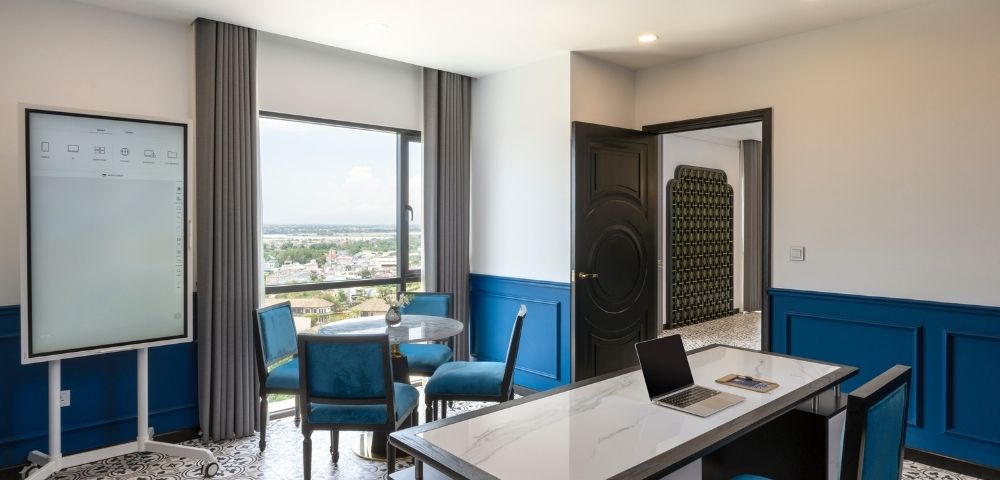 Modern office with a sleek desk, laptop, and four blue chairs around a round table. Large window offers scenic views. Blue accents create a calm atmosphere.
