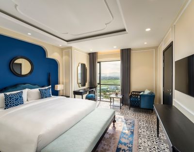 Luxurious hotel room with a large white bed, blue accents, patterned floor, elegant furniture, and a window offering a scenic view. Calm, inviting ambiance.