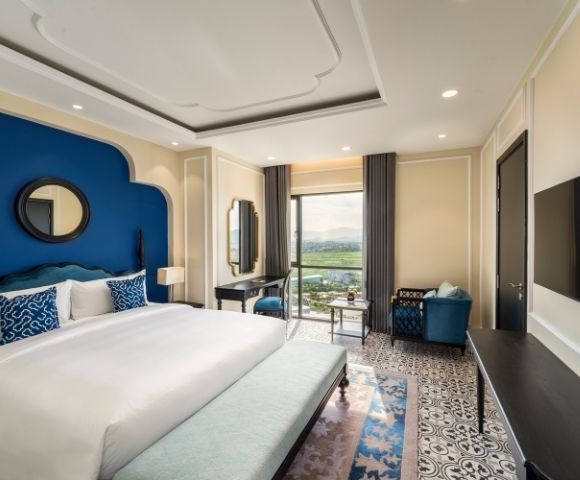 Luxurious hotel room with a large white bed, blue accents, patterned floor, elegant furniture, and a window offering a scenic view. Calm, inviting ambiance.