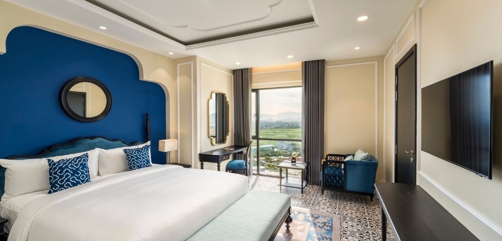 Luxurious hotel room with a large white bed, blue accents, patterned floor, elegant furniture, and a window offering a scenic view. Calm, inviting ambiance.