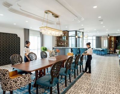 Luxurious dining room with two staff arranging a long wooden table. Elegant chairs, patterned floor, large chandelier, and a modern bar in the background.