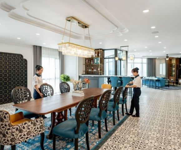 Luxurious dining room with two staff arranging a long wooden table. Elegant chairs, patterned floor, large chandelier, and a modern bar in the background.