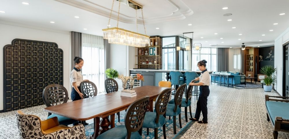 Luxurious dining room with two staff arranging a long wooden table. Elegant chairs, patterned floor, large chandelier, and a modern bar in the background.