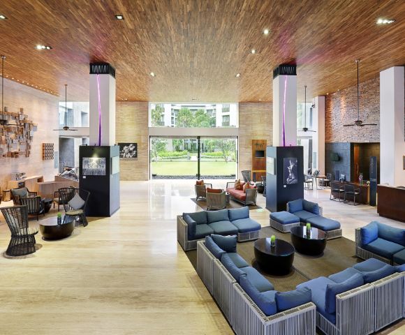 Hotel lobby with modern decor; features blue sofas, wooden ceiling, large window view of greenery, and warm ambient lighting. Airy and welcoming ambiance.