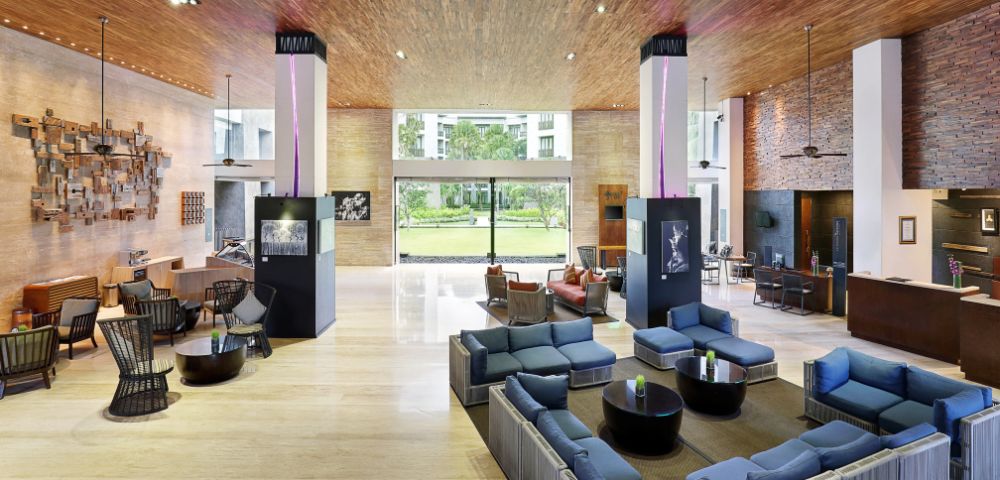 Hotel lobby with modern decor; features blue sofas, wooden ceiling, large window view of greenery, and warm ambient lighting. Airy and welcoming ambiance.