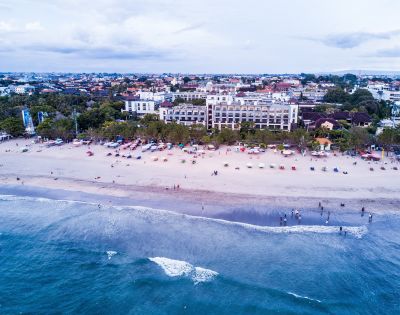 Aerial view of a busy beach with people and colorful umbrellas. Behind the beach, a large hotel and surrounding buildings under a cloudy sky. Serene and vibrant atmosphere.