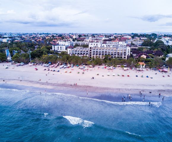 Aerial view of a busy beach with people and colorful umbrellas. Behind the beach, a large hotel and surrounding buildings under a cloudy sky. Serene and vibrant atmosphere.