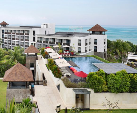Modern resort with white buildings, lush gardens, and a large pool with red umbrellas. Ocean view in the distance under a clear blue sky. Calm and inviting.