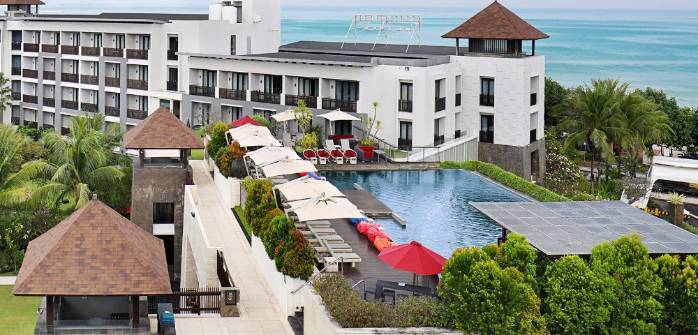 Modern resort with white buildings, lush gardens, and a large pool with red umbrellas. Ocean view in the distance under a clear blue sky. Calm and inviting.