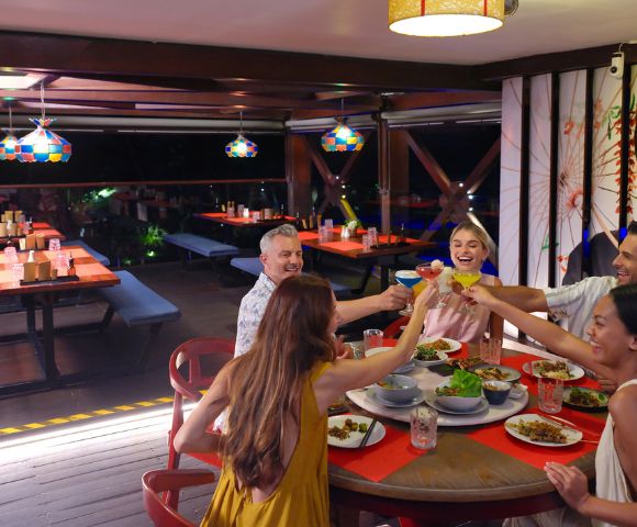 A group of six people sit around a red table in a lively restaurant, joyfully toasting with drinks. Plates of food are spread out in a festive setting.