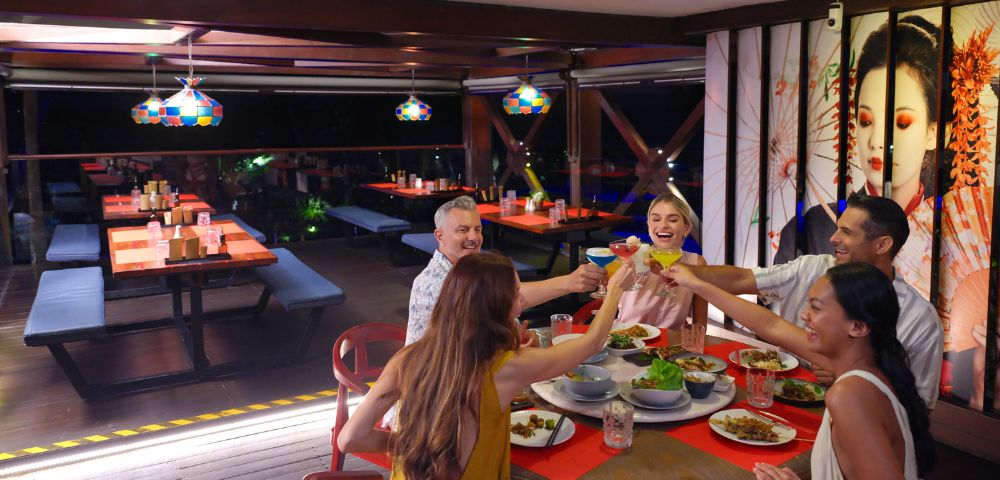 A group of six people sit around a red table in a lively restaurant, joyfully toasting with drinks. Plates of food are spread out in a festive setting.
