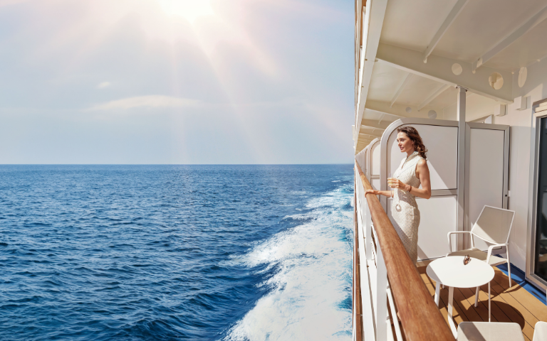 A woman stands on a cruise balcony, enjoying the serene ocean view under bright sunlight, with gentle waves in the distance.