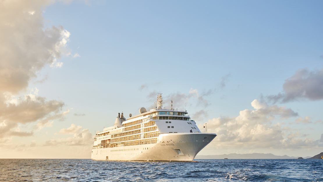A Silversea cruise ship sails through calm waters under a clear sky, surrounded by soft clouds and distant islands.