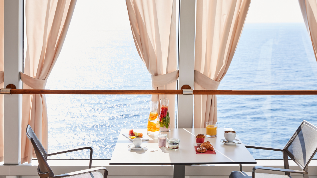 A sunlit table by a sea view, set with a coffee, pastries, fruit, and drinks, surrounded by soft curtains.