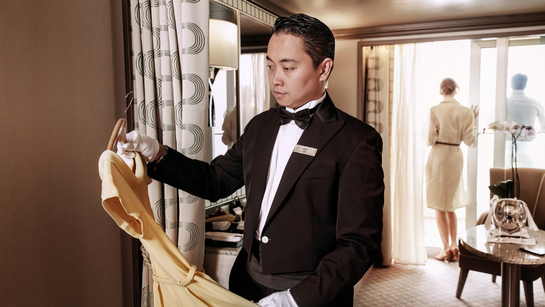 Silversea dedicated butler in formal attire holds a yellow dress, while a woman in a light coat stands in the background, overlooking a balcony.