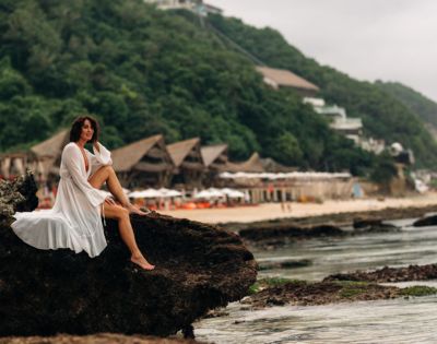 A woman in a flowing white dress sits on a rock by the sea, with lush green hills and beachfront huts in the background, conveying serenity.