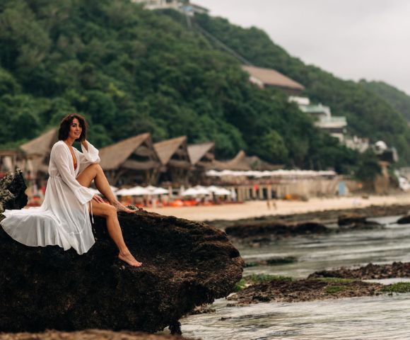 A woman in a flowing white dress sits on a rock by the sea, with lush green hills and beachfront huts in the background, conveying serenity.