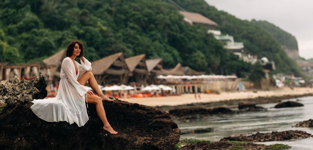 A woman in a flowing white dress sits on a rock by the sea, with lush green hills and beachfront huts in the background, conveying serenity.