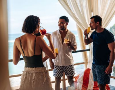 Four friends are enjoying drinks on a balcony by the ocean. They are dressed in summer attire, smiling and chatting, with a relaxed and joyful atmosphere.