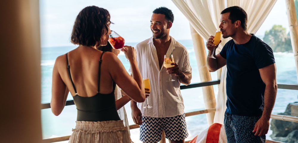 Four friends are enjoying drinks on a balcony by the ocean. They are dressed in summer attire, smiling and chatting, with a relaxed and joyful atmosphere.