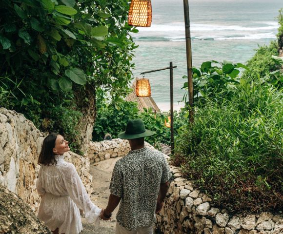 A couple holding hands walks down a stone path, surrounded by lush greenery. Hanging lanterns line the path, leading to a serene ocean view.