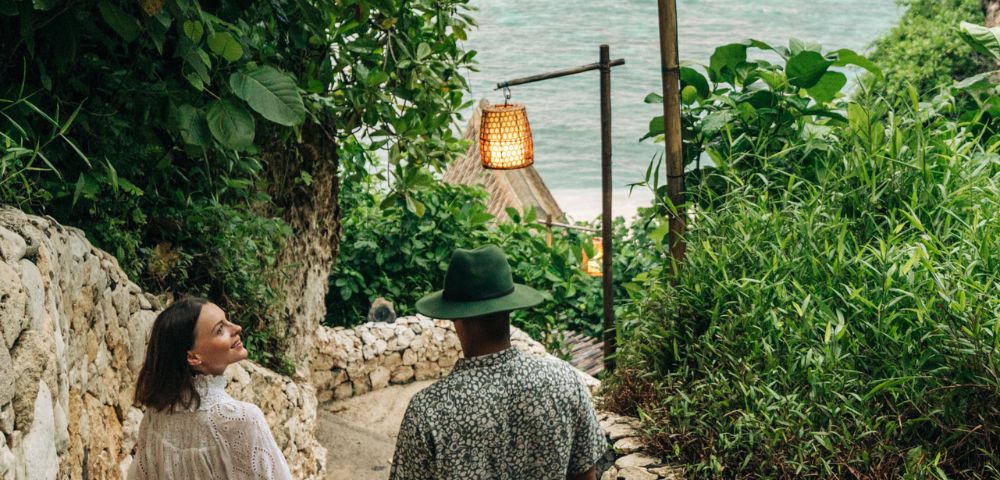 A couple holding hands walks down a stone path, surrounded by lush greenery. Hanging lanterns line the path, leading to a serene ocean view.