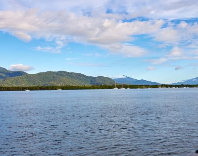 Calm ocean with sailboats in the distance, backed by lush green hills and cloudy blue sky. Serene and peaceful coastal landscape.