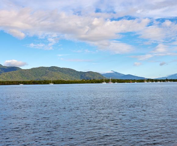 Calm ocean with sailboats in the distance, backed by lush green hills and cloudy blue sky. Serene and peaceful coastal landscape.