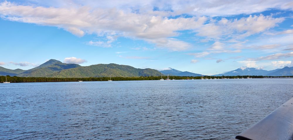 Calm ocean with sailboats in the distance, backed by lush green hills and cloudy blue sky. Serene and peaceful coastal landscape.