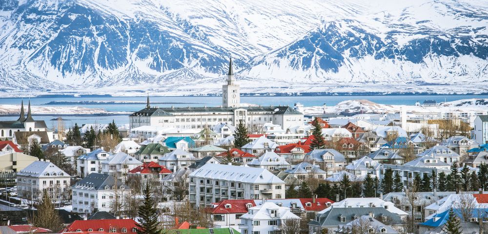 Snow-covered Reykjavik with colorful rooftops, set against majestic mountains and a serene bay. The scene conveys a peaceful, wintery atmosphere.