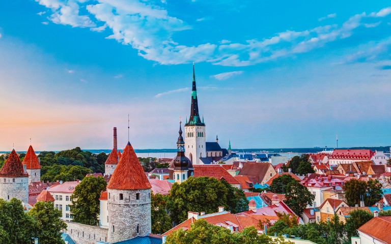 A picturesque view of Tallinn's skyline, featuring medieval towers with red roofs, a tall spire, lush greenery, and a blue sky at sunset. Tranquil and historic.